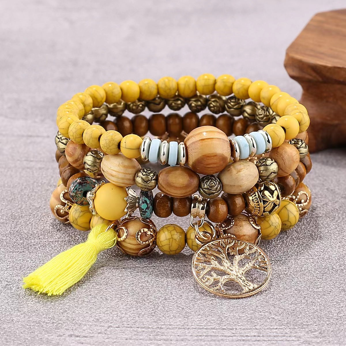 Bracelet à plusieurs rangs de perles jaunes et brunes, avec un pendentif doré arbre de vie et un pompon jaune, posé sur une surface grise.