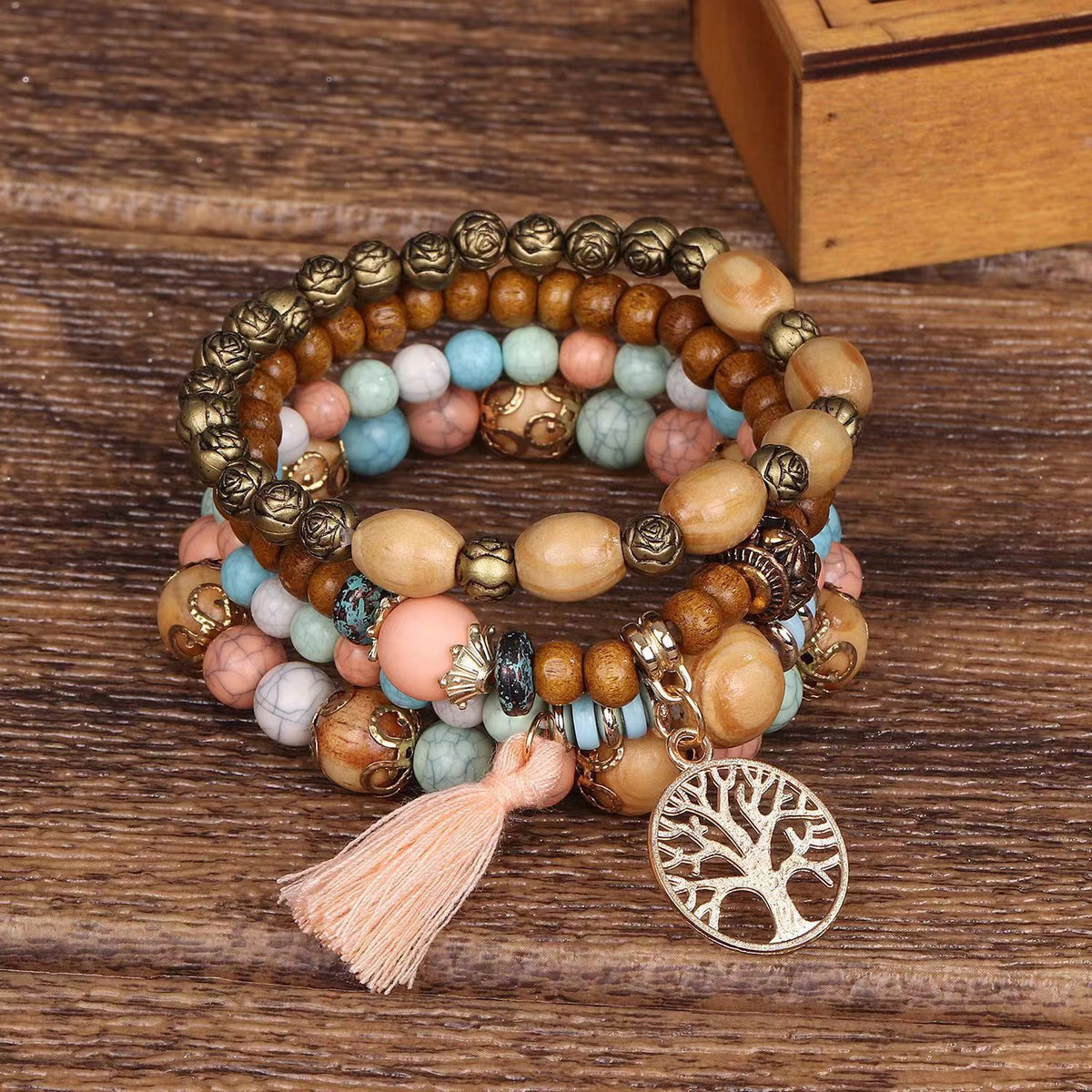 Bracelet à plusieurs rangs de perles multicolores et brunes, avec un pompon rose et un pendentif arbre de vie, posé sur une surface en bois.