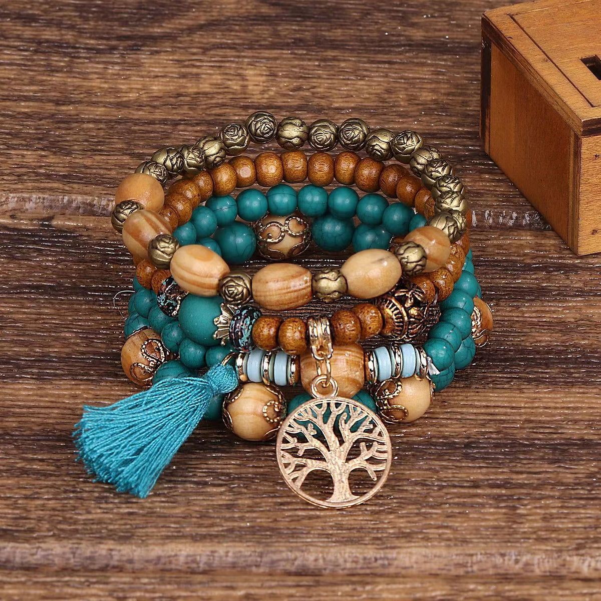 Bracelet à plusieurs rangs de perles turquoise et brunes, avec un pendentif doré arbre de vie et un pompon turquoise, posé sur une surface en bois.