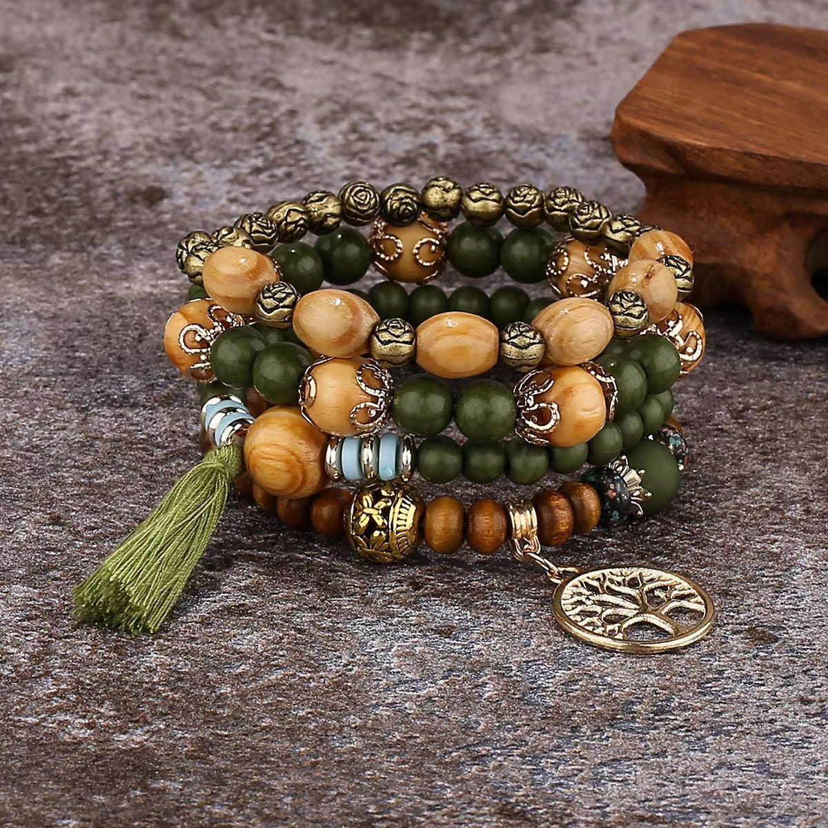Bracelet à plusieurs rangs de perles vert olive et brunes, avec un pendentif doré arbre de vie et un pompon vert olive, posé sur une surface grise texturée.