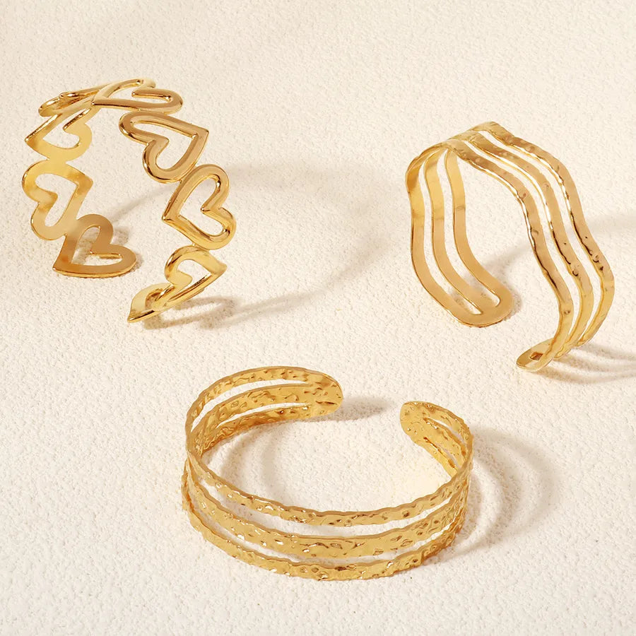 Trois bracelets dorés ouverts : l’un à motifs cœur, l’un à lignes ondulées et l’un à plusieurs rangs texturés, sur fond beige